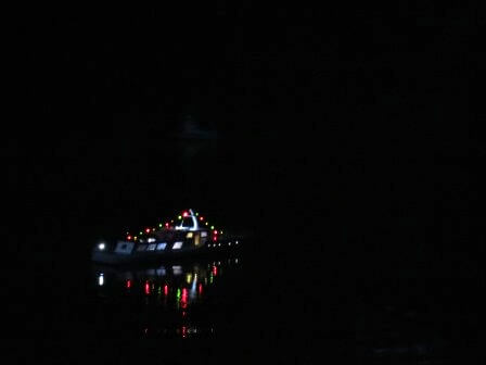Gemütliches Nachtfahren mit einem beleuchteten Schiffsmodell auf ruhigem Wasser, das die Lichter reflektiert.