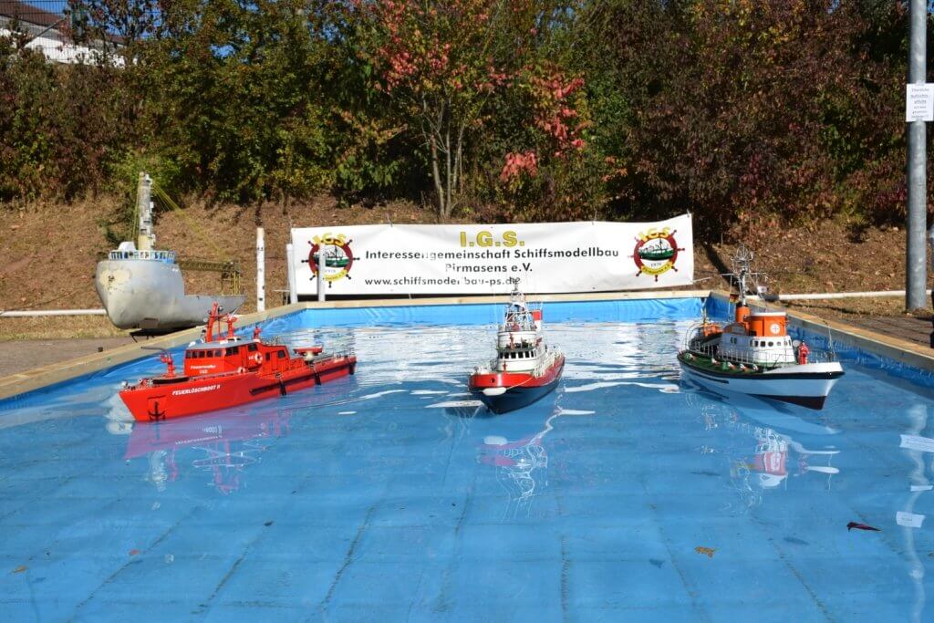 Drei Modellschiffe im Wasser, darunter ein roter Feuerwehrboot und ein grünes Schlepperboot, mit Vereinsbanner im Hintergrund.