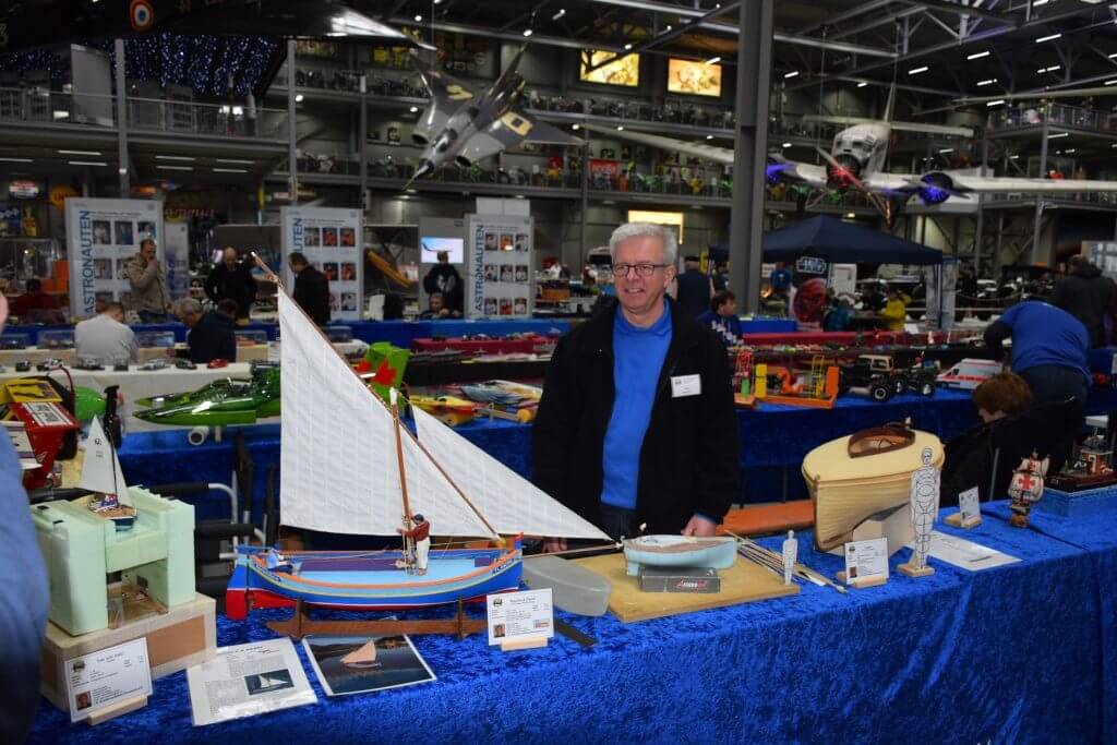 Ein Modellbauer präsentiert seine Schiffsmodelle auf einer Ausstellung, umgeben von verschiedenen Modellbooten und Werkzeugen.