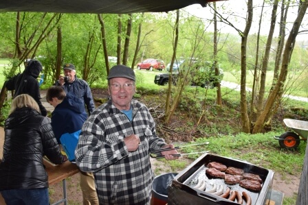 Ein fröhlicher Mann grillt Würstchen und Steaks bei einem Treffen der IG Schiffsmodellbau PS e.V. im Freien.