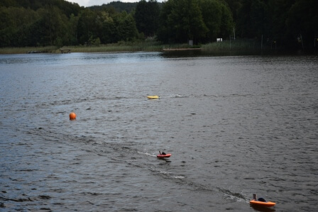 Drei ferngesteuerte Schiffsmodelle fahren auf einem ruhigen See, umgeben von Natur und Bojen.