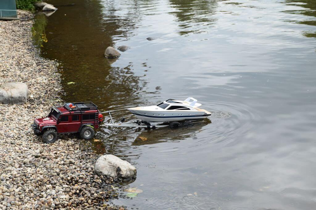 Ein ferngesteuertes Boot wird von einem Modellauto am Ufer eines Gewässers gezogen. Perfekt für Modellbau-Enthusiasten.